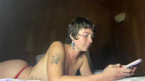 Media: Video of a topless, short-haired woman with tattoos lying on a bed, reading a book. She wears red underwear. The background is dimly lit, wooden walls.