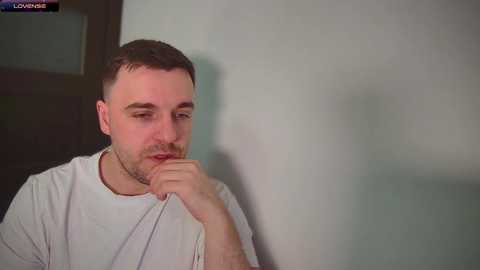 Media: Video of a man with short brown hair, wearing a white T-shirt, sitting against a light grey wall, looking introspective with his hand on his chin.