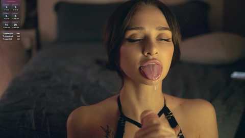 Media: A video of a young woman with dark hair and light skin, wearing a black harness, licking a man's erect penis. The background shows a dimly lit bedroom with a bed.