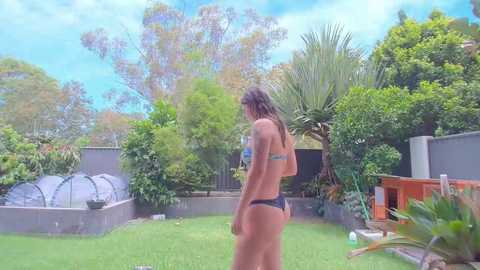 Media: Video of a young woman in a bikini, with long, wet hair, standing on a lush green lawn in a backyard. The background features a variety of greenery, a small pool, and a shed.