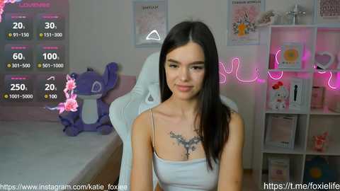 Media: A video of a young woman with fair skin, long dark hair, and a tattoo on her chest, sitting on a white chair in a brightly lit room with pink neon lights and decorations.