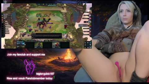 Media: Video of a woman in a brown jacket, blonde hair, and pink dildo, lying on a bed with a TV showing a League of Legends game, background featuring a fire.