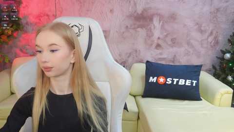 Media: Video of a young, slim, fair-skinned woman with long blonde hair wearing a black top, sitting on a white gaming chair. Background features a pink textured wall, a yellow couch, and a \"MOSTBET\" pillow.