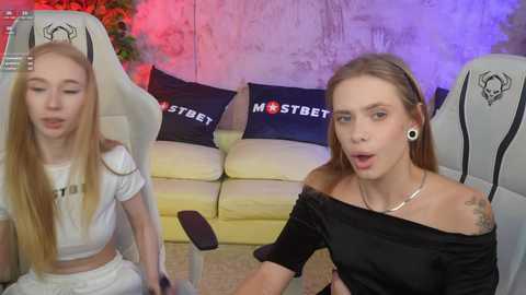 Media: Video of two young women in off-shoulder black tops, sitting in gaming chairs with \"MasterBet\" pillows, in a room with a colorful, industrial backdrop.