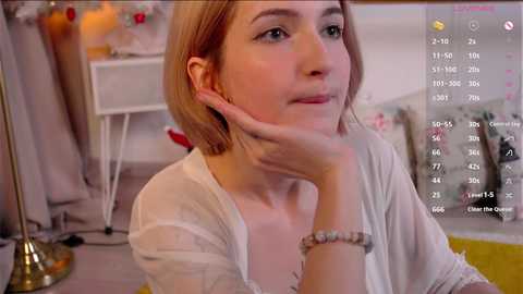 Media: Video of a fair-skinned, blonde woman with a white sheer top, sitting pensively with her chin on her hand. Background shows a cozy, brightly lit room with white furniture and a calendar overlay.