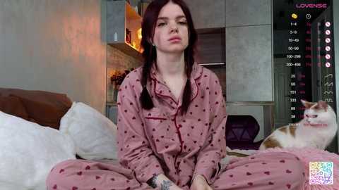 Media: Video of a young Caucasian woman with long, dark hair in pink pajamas, sitting cross-legged on a bed with a white cat. The room features modern decor, with a fireplace in the background.