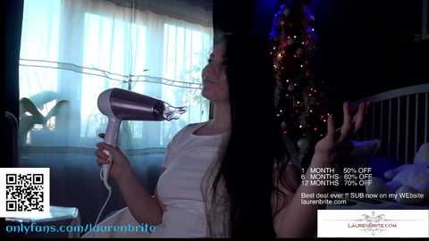 Media: Video of a woman with long dark hair using a hairdryer in a dimly lit room with a Christmas tree and a bed in the background.