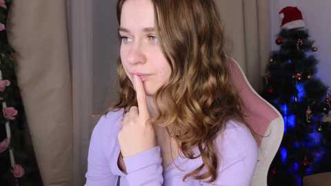 Media: Video of a young Caucasian woman with wavy, light brown hair, wearing a light purple top, sitting in a pink gaming chair. She has her index finger to her lips in a shushing gesture. Background includes a Christmas tree with decorations.