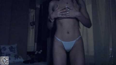 Media: A dimly lit video of a slender, topless woman with fair skin, wearing white panties, covering her breasts with her hands. The background features a dimly lit room with a floral pillow and a door.