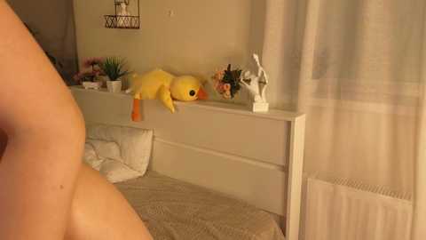 Video of a cozy bedroom with a white bed, yellow plush duck toy, white curtains, and a small decorative figure on a shelf. Warm lighting creates a soft, inviting atmosphere.