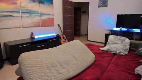 Media: Video of a cozy, modern bedroom with a beige sofa, a dark wooden dresser, a guitar, a TV, and a wall painting of a beach scene.