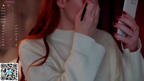 Media: Video of a fair-skinned woman with long red hair, wearing a white sweater, speaking on a phone, with a blurred background and a QR code in the bottom left corner.