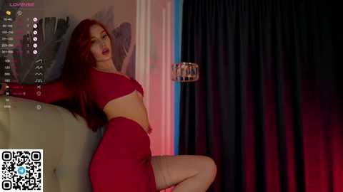 Media: Video of a curvy woman with long red hair, wearing a red crop top and matching skirt, leaning against a beige couch. Dark curtains and a modern light fixture in the background.