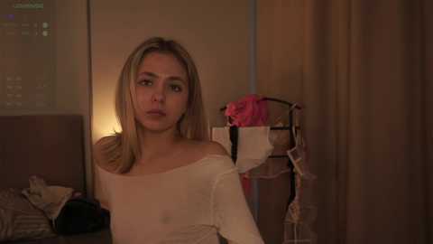 Media: A video shows a young, blonde woman with a solemn expression in a dimly lit bedroom. She wears a sheer, off-shoulder top. The background includes a bed, a pink garment, and a metal rack.