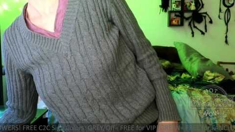 Media: Video of a woman in a gray ribbed sweater, leaning against a bed with green sheets and a patterned blanket. The background features a wall adorned with a black chandelier and framed photos.