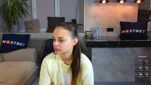 Media: Video of a young woman with long brown hair in a ponytail, wearing a yellow shirt, sitting indoors near a couch with dark blue cushions. Modern living room with gray walls, plants, and a black marble countertop.