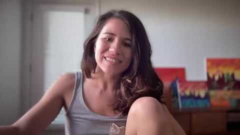 Media: Video of a smiling young woman with long brown hair, wearing a gray tank top, sitting in a brightly lit room with colorful paintings on the wall and a white door in the background.