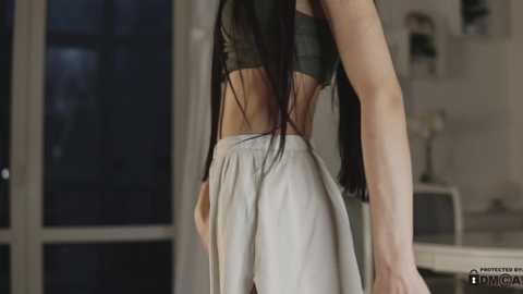 Media: A video of a slender woman with long black hair, wearing a dark green crop top and white pants, standing indoors with a blurred background of a modern room with large windows and a bookshelf.