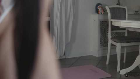 Media: Video of a woman with long black hair, wearing a black top, standing in a modern dining room with white furniture, a blue decorative item, and a gray tiled floor.