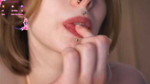 Media: Video of a close-up of a light-skinned woman with fair skin, wearing a silver nose ring and small earrings. She has a hand raised to her mouth with a playful expression. Background is blurred.