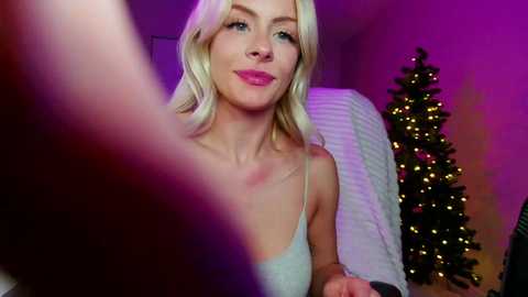 Media: Video of a smiling blonde woman in a gray tank top, sitting in a room with a lit Christmas tree in the background.