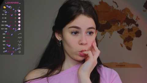 Video of a young woman with long black hair, pale skin, and a pensive expression, resting her chin on her hand. She wears a light pink off-shoulder top. Background features a digital map and social media icons.