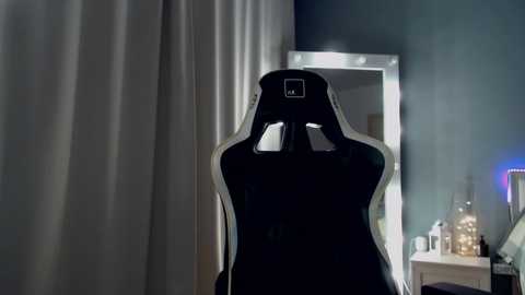 Media: Video of a sleek, black gaming chair with white accents and a small, rectangular camera on its headrest, situated in a dimly lit room with a large, silver-framed mirror and a desk with scattered objects.