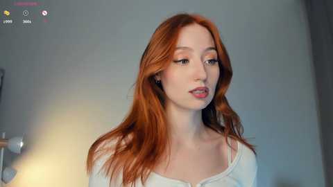 Media: Video of a young woman with long, straight red hair, pale skin, and a slender figure, wearing a white off-shoulder top. She has a serene expression. Background is a minimalist, softly lit room with a light gray wall.