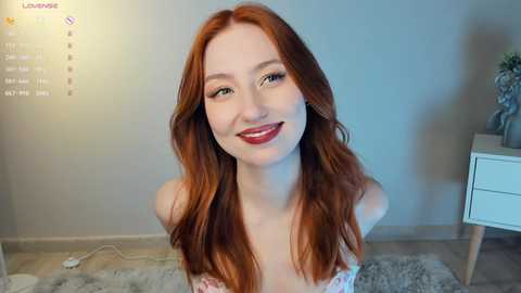 Media: Video of a young, fair-skinned woman with long, wavy red hair, smiling with red lipstick, wearing a strapless pink top, standing in a softly lit room with a white dresser and a fluffy gray rug.