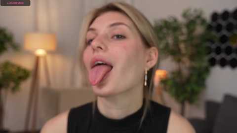 Media: A video of a blonde woman with light skin, sticking out her tongue in a playful manner. She wears a black top and has dangling earrings. Background features a lamp, green plants, and a black wall with circular decorations.