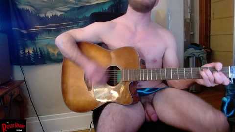 Media: Video of a shirtless, pale-skinned man with a trimmed beard playing a wooden acoustic guitar in a cozy room with a tapestry of mountains, wooden floor, and a door.