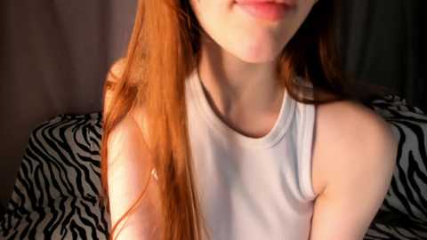 Video of a fair-skinned woman with long, straight red hair, wearing a white sleeveless top, against a backdrop of zebra-print fabric.