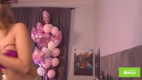 Video of a woman in a pink bra, holding a bunch of pink balloons in a room with a gray curtain and a framed picture of a lake.