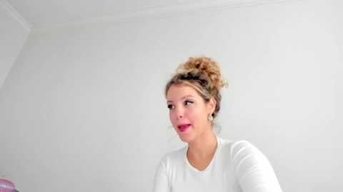 Media: A video of a young woman with light skin and blonde hair in a curly updo, wearing a white top, smiling against a plain white wall background.