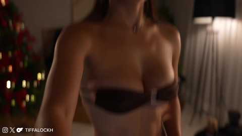 Media: Video of a woman with medium breasts, wearing a strapless black bra, in a dimly lit, cozy room with a Christmas tree and a lamp in the background.