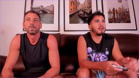 Media: Video of two muscular men, one with a beard, sitting on a brown leather couch. They wear black sleeveless shirts and are watching a video game. Background features framed Venice canal photos.