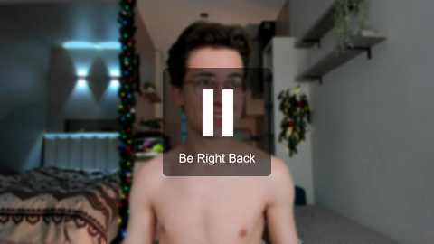 Media: Video of a shirtless young man with short brown hair, blurred face, in a dimly lit bedroom with a bed and festive decorations. Text overlay: \"Be Right Back.\