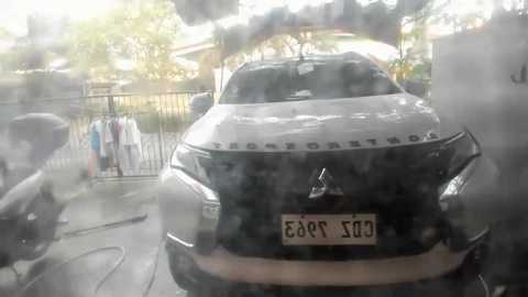 Media: A video captures a black Mercedes-Benz car with license plate \"\u0395\u0391 1000\" obscured by smoke and steam. The background shows a city street, a fence, and a person in casual clothes. The scene appears to be a car wash.