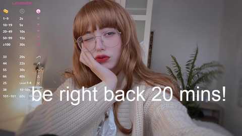 Media: Video of a pale-skinned, red-haired woman with glasses and bangs, wearing a cream sweater, resting her chin on her hand, surrounded by green plants. Text overlay: \"be right back 20 mins!\