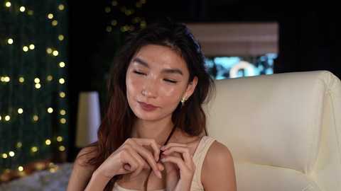 Media: Video of an Asian woman with long, dark hair, wearing a beige sleeveless top, adjusting her necklace in a dimly lit room with Christmas lights and a fireplace in the background.