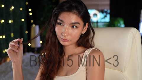 Media: Video of a young woman with long, wavy dark hair, fair skin, and brown eyes, wearing a beige tank top, seated indoors with a blurred background.