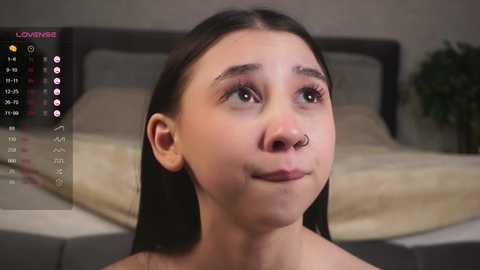 Media: Video of a young, light-skinned woman with straight black hair, a nose ring, and a small smile, standing in a bedroom with a gray bed and a calendar displaying her daily schedule.