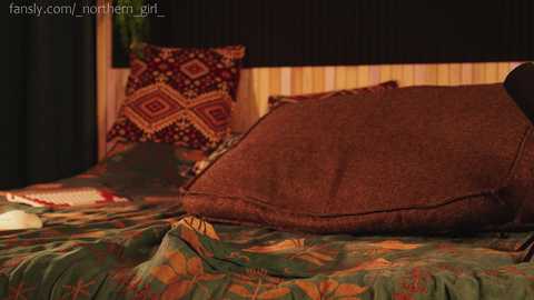 Media: Video of a cozy, dimly lit bedroom featuring a bed with a green, leaf-patterned duvet, a large brown pillow, and a decorative pillow with a red and gold geometric design. The background includes vertical wooden blinds.