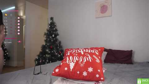 A video of a woman in a light pink dress standing next to a Christmas tree and a bed with red pillows and a \"Merry Christmas\" pillow.