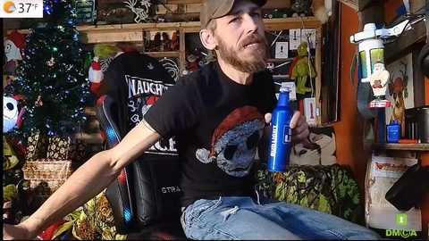 Media: Video of a bearded, fit man in a black \"Naughty Santa\" shirt, jeans, and cap, sitting on a gaming chair, holding a blue water bottle. Background features a decorated Christmas tree, shelves with toys, and a chaotic room.