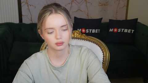 Media: Video of a young, fair-skinned woman with blonde hair tied back, wearing a light green shirt, sitting on a green velvet couch with gold trim. Background features a floral wallpaper and two black pillows with white \"MOSTBET\" logos.