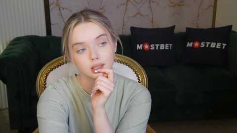 Media: Video of a young woman with light skin and blonde hair, wearing a green top, sitting on a dark green couch with black pillows, looking thoughtful.