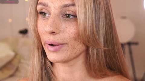 Media: Video of a fair-skinned woman with long, blonde hair, green eyes, and freckles, wearing subtle makeup, smiling slightly, in a softly lit room.