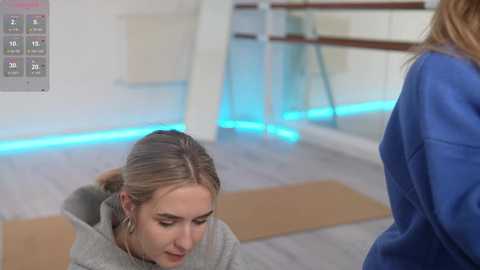 Media: Video of a blonde woman in a gray sweater meditating on a yoga mat in a modern studio with blue LED lighting, a blue-clad figure standing, and ballet barre in background.