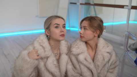 Media: Video of two women in fur coats, one blonde with dark eyebrows, the other brunette, in a modern, minimalist room with neon blue lights and mirrored walls.